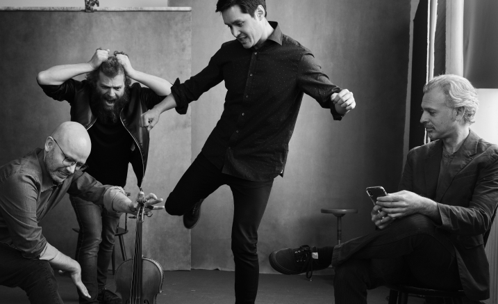 Brooklyn Rider. A Black and white image of four men clowning. One has his arms out and one leg in the air, the other three sit around him, one gesticulating with his viola, one with his hands on his head and the third siting quite demurely with his hands holding an iPhone.