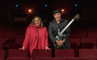Members of First Nations music duo, Gina Williams (left) and Guy Ghouse (right). Williams is a woman with long brown and blonde hair, light brown skin, a big smile and wearing a red dress with the same coloured scarf. Ghouse is a tall man with light brown skin, wearing a newsboy hat and holding a black electronic guitar, wearing a black shirt and smiling.
