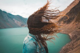 A woman in a denim jack looks out over a mountain lake. We cannot see her face: the focus of the photo is on her long red hair blowing in the wind.