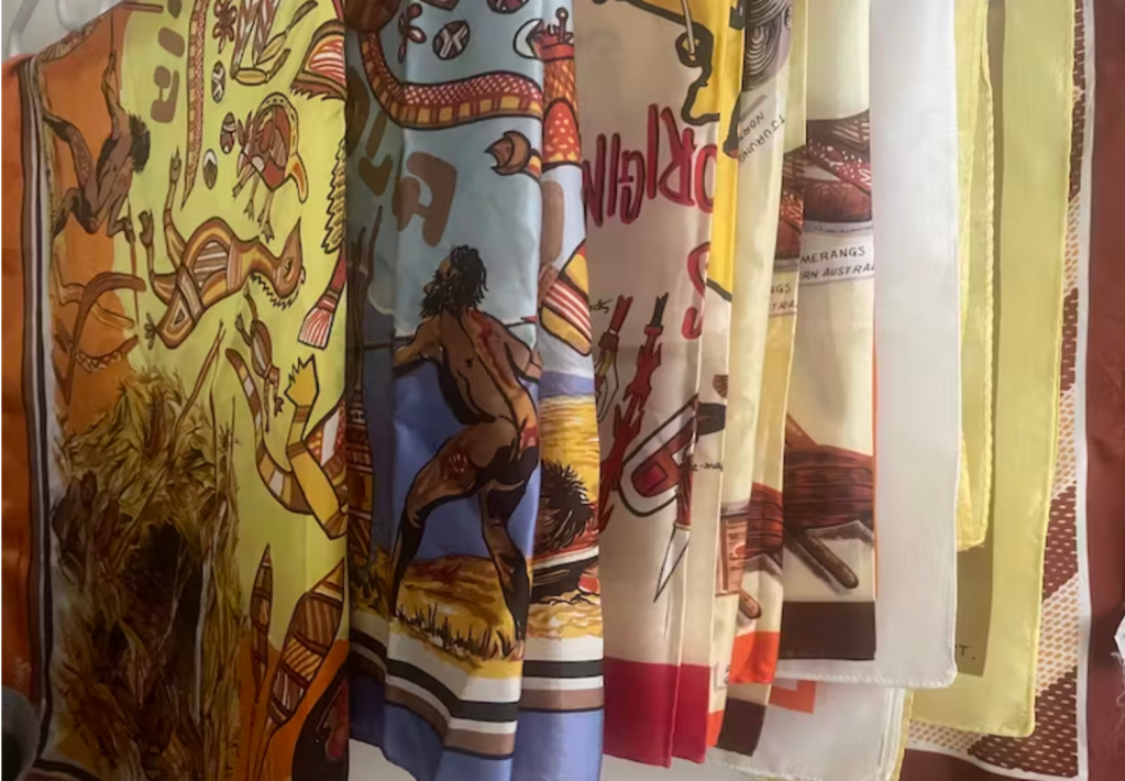 First Nations garments. Image is a row of scarves hanging down and depicting kitsch images of Aboriginal people and culture.