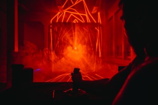 Hyperbolic Psychedelic Mind Melting Tunnel of Light, the image is the silhouette of a person holding a joystick in the foreground and behind them an explosion of red laser lights.