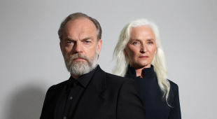 Two white people in back clothes stand against a grey background. The man has a receding hairline and a short beard; the woman has long white hair.