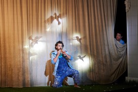The Magic Flute. A man in blue overalls and shirt playing a flute. There are silhouettes of birds on a curtain behind him.