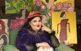 A women dressed in 1940s fashion with rosy cheeks and a thoughtful gaze. She is wearing a purple velvet dress, with black feathered cuffs, holding a handbag shaped like a fan and a red beret. She sits on a worn green armchair and behind her are several paintings, including self portraits with a gold background.