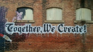 Community engaged. Image is a brick wall with three arched windows at the top filled in with corrugated iron. There is a frame wide mural on the bricks that says Together we Create!