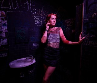 overflow. A woman stands next to a basin in a graffitied club toilet. She is wearing a fishnet top and short black skirt and preening for the camera.
