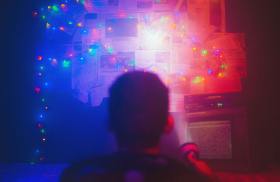 Photo: Matheus Bertelli, Pexel. A boy is pointing a flashlight onto a board stuck with newspaper snippets in a dark room. There are fairy lights across the board, emitting a blue and red hue.