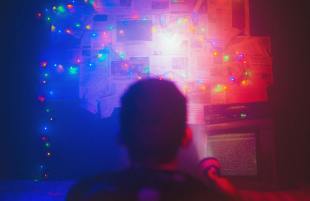 Photo: Matheus Bertelli, Pexel. A boy is pointing a flashlight onto a board stuck with newspaper snippets in a dark room. There are fairy lights across the board, emitting a blue and red hue.