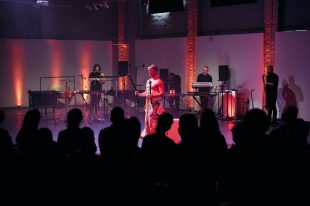 Portent. A group of musicians and actors perform a ritual inspired by eclipses. Silhouettes of the audience are at the front of the shot.