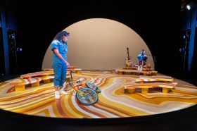 The Hate Race. A woman in blue is standing on a stage marked with yellow and brown wavy patterns. She next to a bicycle and there is a large half circle light shape on the back wall.