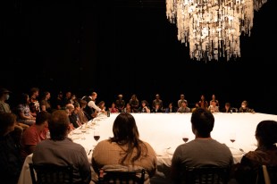 ‘Food’ at Perth Festival 2024. Photo: Maria Baranova. Audiences sit at a large table covered by a white table cloth. A glowing chandelier hangs above them, while the performer is serving audiences wine.