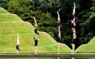 A series of acrobats perform handstands, two-highs and three-highs by the edge of a body of water, in which their reflections are visible.