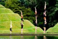 A series of acrobats perform handstands, two-highs and three-highs by the edge of a body of water, in which their reflections are visible.