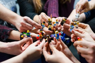 Photo: Vlad Hilitanu, Unsplash. Hands holding LEGO figures. They are all pointing their figures towards the centre.
