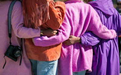 Photo: Vonecia Carswell, Unsplash. Four women hugging each other from behind their backs, wearing different shades of purple clothing representing International Women's Day.