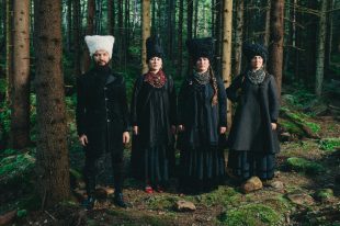 Four people stand in a forest wearing long black coats. The three women also wear tall black hats, the man a tall white one.