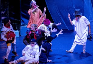 The Great Travelling Médecin Show. A range of actors in various eclectic costumes are variously standing and sitting on the stage reaching out toward an actor in a white tunic with a top hat and a doctor's bag.
