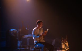 Artist and event presenter registrations open for Sydney Fringe 2024. Photo: Supplied. A White man with short brown hair sitting on a stage next to a simple bed. Stage light is shining on him and he is holding a small book, looking slightly above with his chin tilted.