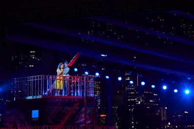 West Side Story by Opera Australia. A dark harbourside long shot of two young lovers - Tony and Maria - standing on metal balcony embracing, with a cityscape behind them.