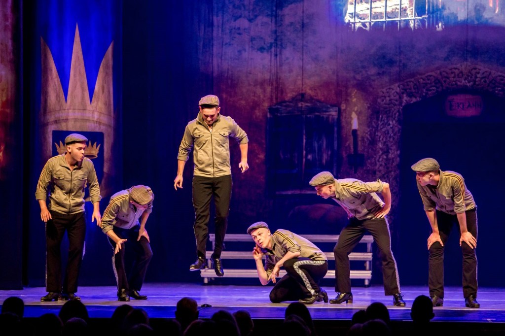 Sox men perform on a stage lit in blues and purples. They wear traditional Irish caps, dark trousers and tan-coloured caps. One leaps into the air exuberantly; the other five lean in to listen to his percussive footwork when he lands.