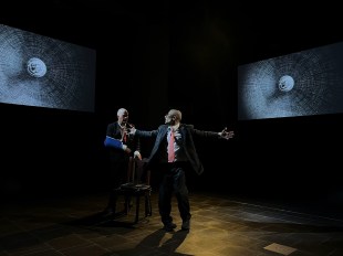 Two of Them. Two men in business suits on a stage. One is in the centre with his arms outstretched to the side, the other sitting behind on a stool with his arm in a sling. There are two abstract projections of spheres on the back wall.