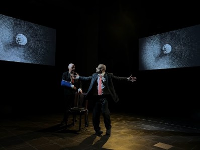 Two of Them. Two men in business suits on a stage. One is in the centre with his arms outstretched to the side, the other sitting behind on a stool with his arm in a sling. There are two abstract projections of spheres on the back wall.