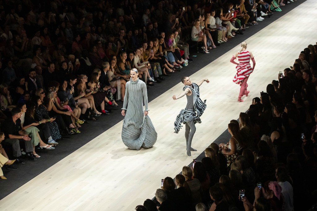 Designs by Alexander Enticknap at the Paypal Melbourne Fashion Festival 2024 National Graduate Showcase. Photo: Lucas Dawson. A view of the runway with three models taken from a high vantage point. The models are wearing garments that feature exaggerated forms in black and red stripes. A crowd is sitting on both sides of the runway.