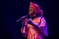Angélique Kidjo. A woman of African appearance in a red patterned African dress and turban sings into a microphone against a black backdrop.
