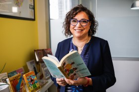 A middle-aged woan with dark neck length wavy hair and dark rimmed glasses wears a dark shirt and jacket, smiles at the camera and holds a book. She is standing in a room with more books on a ledge next to her. Opportunities and awards.