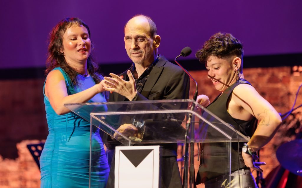 L to R: Autumn Skuthorpe, Brenden Borellini and Georgia Cranko. Photo: Luke Monsour, Bulimba Studio. Three people stand on a stage behind a clear podium at the Matilda Awards. Skuthorpe has long curly brown hair, rosy cheeks, and an aqua-coloured dress. Skuthorpe is writing on the palm of Borellini, who has closely shaved hair, short grey stubble and wearing a black suit. Cranko has short curly brown hair, wearing a black vest and grey pants.