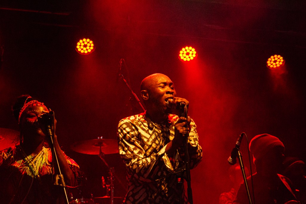On a red and orange spotlit stage a man with a bald head and patterned shirt cradles a mic and sings passionately.