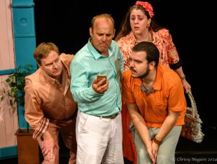 The Government Inspector. Three men in brightly coloured shirts look at the phone the middle one is holding. A woman behind is peering over his shoulder to see too. They all look concerned.