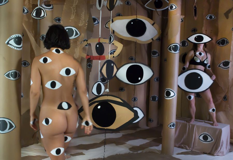 Women's bodies in art: a photograph of a naked woman with her back to the camera surrounding by many paper cut out illustrations of eyes and carboard assemblages.