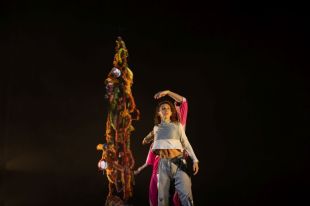 A female dancer in white and grey costume stands in front of a colourful stringy sculpture against a black background.