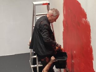 Elderly man is sitting on a step ladder in a black suit with his eyes closed. He is dipping a paint brush into a tub held by another person and on the wall is a large splash of red paint.