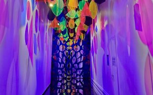 ‘Joy’, installation view at Immigration Museum. Photo: ArtsHub. A corridor with neon plastic mobiles hanging overhead that reflect light onto both sides of the walls. The space is lit in purple light with more shaped mirrors at the end of the corridor.