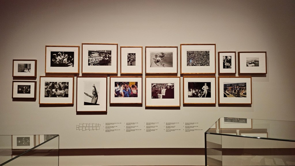 Photographs dating from 1968 to 1983 in ‘Melbourne Out Loud: Life through the lens of Rennie Ellis’ at State Library Victoria. Photo: ArtsHub. 15 photographs hung in two rows on a white gallery wall, depicting scenes from protests on the streets to a family at the pool. 