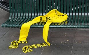 ‘Safety Yellow Woman’ by Sophia Cai. Photo: Supplied, courtesy the artist. A bright yellow sweater with ridiculously long sleeves and black stripes is laid out on a green metal bench. Cai speaks on the cost of her craft.