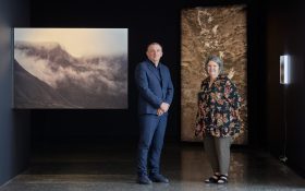 MAMA CEO Blair French and 2024 National Photography Prize guest judge Nici Cumpston standing next to ‘Four Days Before Winter’, a series of work by Ellen Dahl. Photo: Jeremy Weihrauch. The photos are displayed in varying scales in a dim gallery space, showing flowing hills.