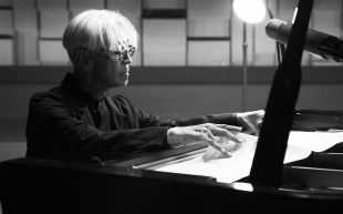 Still from ‘Ryuichi Sakamoto | Opus’. Image: Supplied. A black and white image of Sakamoto, a Japanese man in his 70s with white hair, tortoiseshell glasses and a black blazer at the piano. He is sifting through some sheet music.