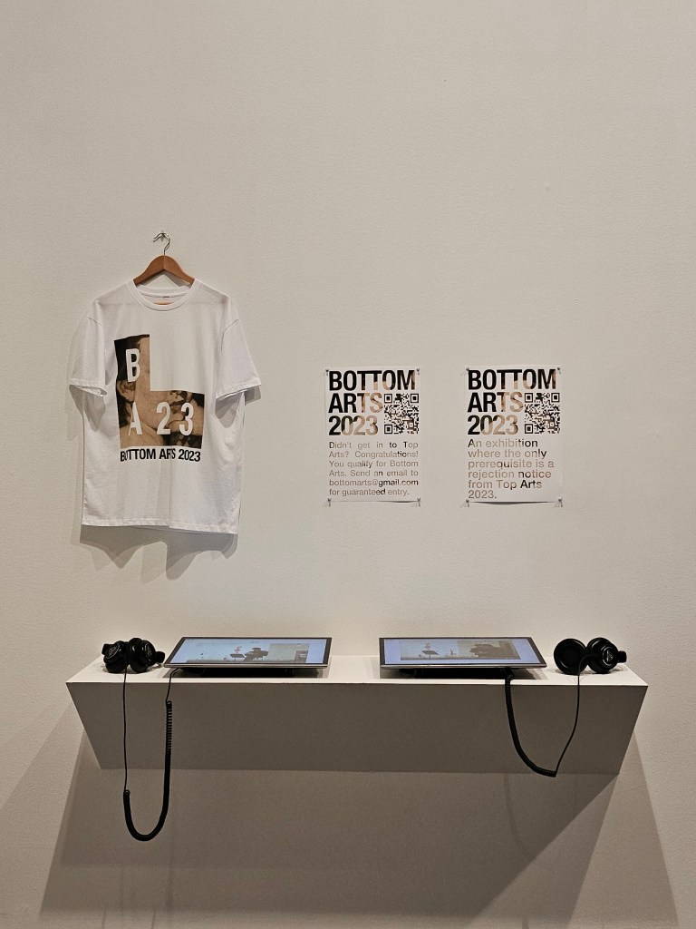 Elliot Broome, ‘Bottom Arts 2023’, installation view at ‘Top Arts 2024’. Photo: ArtsHub. Two posters with the text ‘BOTTOM ARTS 2023’ and a QR code’ are pinned on a white wall. To its left is a t-shirt printed with the ‘BA23’ logo. Beneath them are two screens with headphones. 