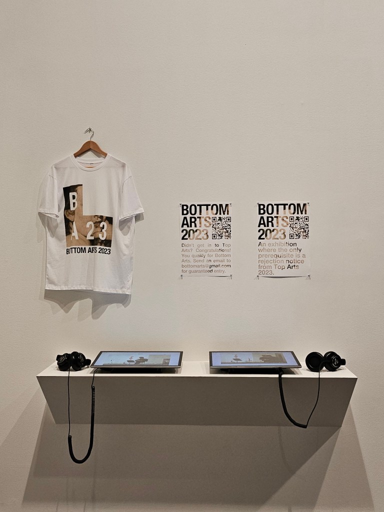 Elliot Broome, ‘Bottom Arts 2023’, installation view at ‘Top Arts 2024’. Photo: ArtsHub. Two posters with the text ‘BOTTOM ARTS 2023’ and a QR code’ are pinned on a white wall. To its left is a t-shirt printed with the ‘BA23’ logo. Beneath them are two screens with headphones. 