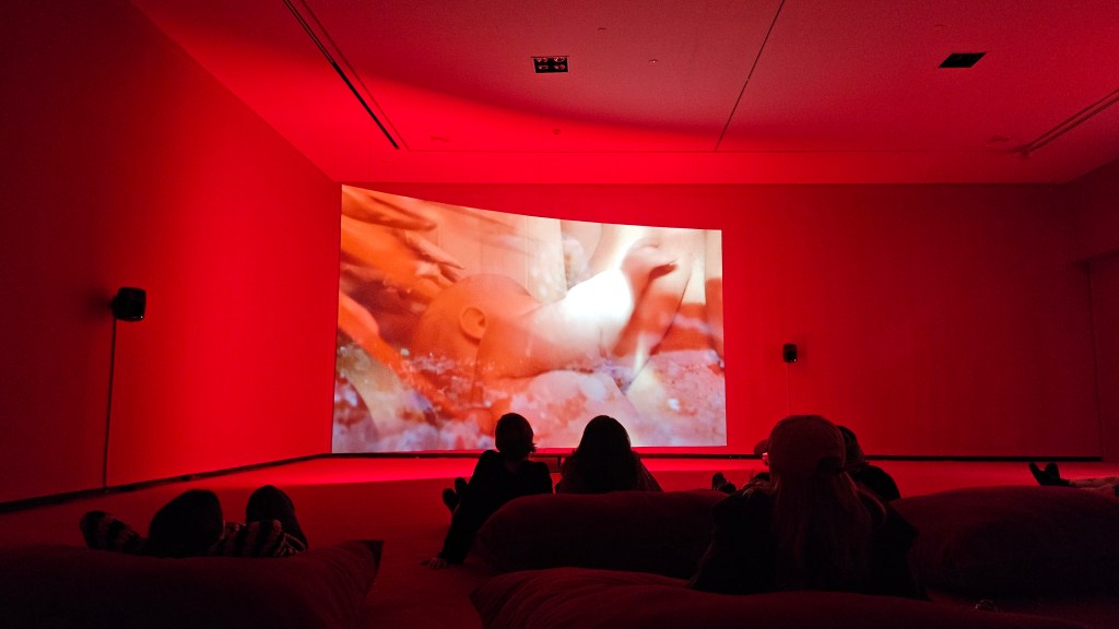 Laure Prouvost, ‘Four for see beauties’, 2022, installation view at ACCA. Photo: ArtsHub. Viewers sitting on beanbags in a red room with a large screen in one corner. On the screen is a small infant being fed on the breast. 