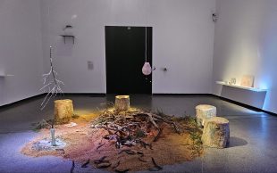 Laure Prouvost, ‘Gathering Ho Ma, The glaneuse’, 2023, installation view at ACCA. Photo: ArtsHub. An installation of many components include a makeshift camp with red dirt and branches, seats, a hanging lightbulb shaped like a breast.