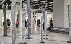 Naminapu Maymuru-White, ‘Larrakitj Forest’, 2024, presented by Sullivan+Strumpf as part of Encounters, Art Basel Hong Kong 2024. Photo: ArtsHub. A dozen organic wood poles with black and white dotted paintings inside the convention centre.