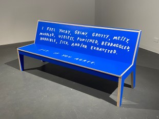 Blue bench with white text painted on it in a gallery setting.
