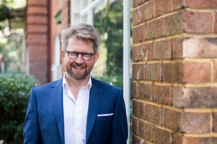 Iain Grandage. Smiling man with fair hair, glasses and beard, in white shirt and blue jacket, outside leaning against a brick wall, smiling at camera.