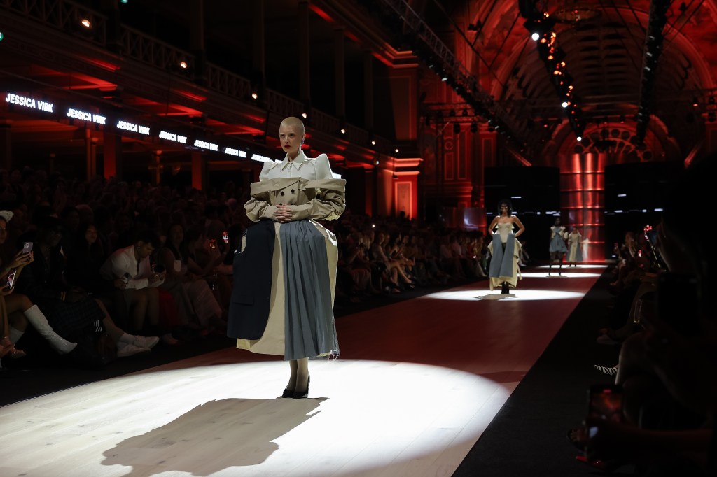 Designs by Jessica Virk at the Paypal Melbourne Fashion Festival 2024 National Graduate Showcase. Photo: Lucas Dawson. Four models standing under a spotlight on the runway with red lights illuminating the space. They are wearing garments deconstructed from trench coats in gray and camel.