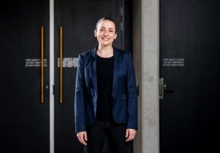 A white woman dressed in black with a dark blue jacket stands outside a theatre door. Her short hair is pulled back and she is smiling at the camera.