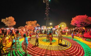 Parrtjima 2023. Photo: Lisa Hatz. People gathering at the Parrtjima festival with kids and an adult at the centre of a vibrnat light project, surrounded by trees and the natural landscape.